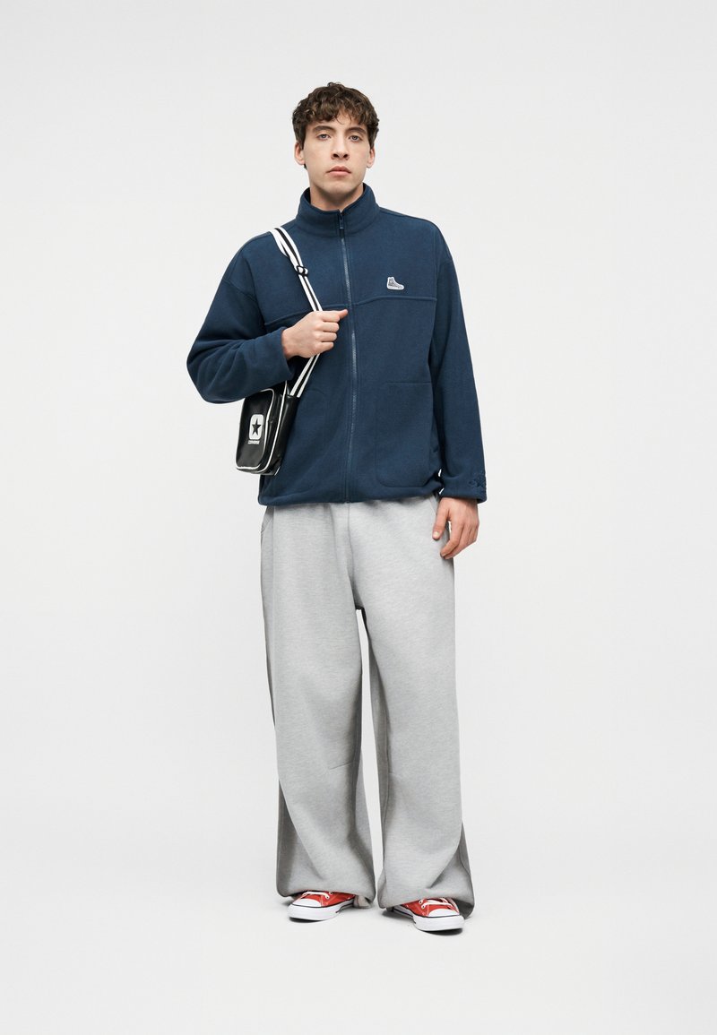 Young man wearing navy zip-up jacket, gray wide-leg pants, red sneakers, and a black crossbody bag with white star and stripes.