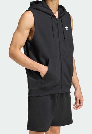 Sweat à capuche noir sans manches zippé et short assorti porté par une personne avec une main dans la poche du sweat, montrant une tenue sportive décontractée.