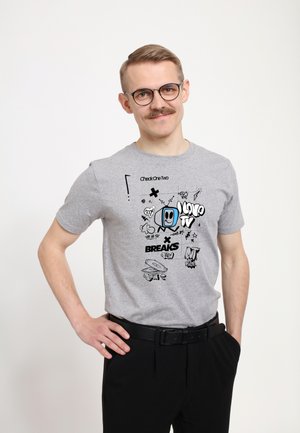 Mann mit Brille und Schnurrbart, trägt ein graues T-Shirt mit Cartoon-Fernseher und Text „Breaks“ sowie schwarze Hose, steht mit einer Hand in der Hüfte.