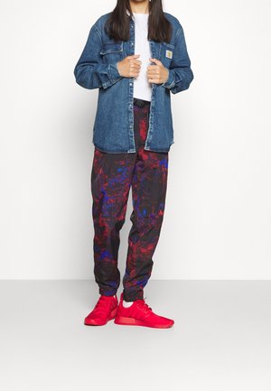 Personne portant une veste en jean bleue sur une chemise blanche, un pantalon à motifs multicolores et des baskets rouges vives, debout contre un mur blanc.