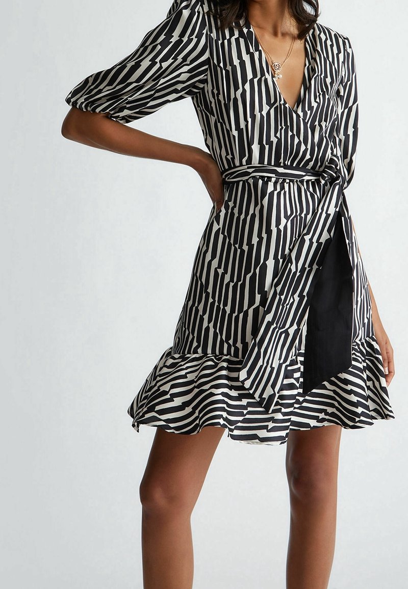 Femme portant une robe portefeuille à rayures noires et blanches avec des manches bouffantes, un ourlet à volants et une ceinture nouée à la taille, affichant une longueur mi-cuisse.