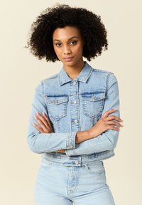 Chaqueta de mezclilla azul claro con un diseño recortado, dos bolsillos en el pecho, cierre de botón y cuello clásico. Llevada sobre jeans azul claro.