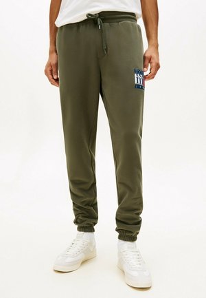 Person trägt olivgrüne Jogginghosen mit elastischen Bündchen und einem Logo am Oberschenkel, kombiniert mit weißen Sneakers und einem weißen T-Shirt.