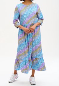 Žena v modrých šatech s diagonálními pastelovými srdíčky a bílých teniskách s růžovými akcenty, stojící se sepnutýma rukama.