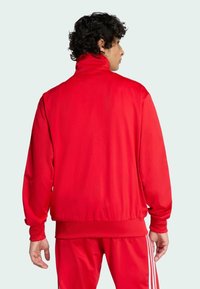 Crvena atletska jakna s visokim ovratnikom i elastičnim mansetama. Glatka, sjajna tkanina s opuštenim krojem, u kombinaciji s crvenim hlačama i bijelim prugama.