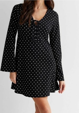 Kvinde iført en sort, langærmet, prikket kjole med nøglehul-udskæring bundet med en sløjfe, stående mod en ensfarvet baggrund.