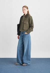 Afgekorte olijfgroene jas met een bruine kraag en voorzakken, gecombineerd met wijde blauwe jeans en grijze sneakers. Eenvoudige zwarte totebag.