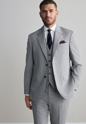 Mann in einem hellgrauen Dreiteiler mit weißem Hemd, dunkel gestreifter Krawatte und bordeauxrotem Einstecktuch, steht mit einer Hand in der Tasche.