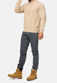 Beige gebreide trui met een hoge kraag en een drieknops sluiting, gecombineerd met grijze slim-fit jeans en tan veterlaarzen. Duurzame materialen.