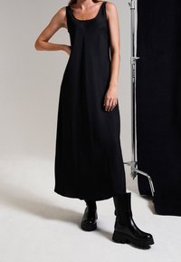 Mujer vistiendo un vestido midi negro sin mangas y botas de plataforma gruesa negras, de pie con una mano en la cadera frente a un fondo liso.