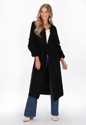 Schwarze, gestrickte lange Strickjacke mit strukturierten Ärmeln, offenem Vorderteil und gebundenem Bund. Getragen über einem weißen Oberteil und kombiniert mit weit geschnittenen Jeans.