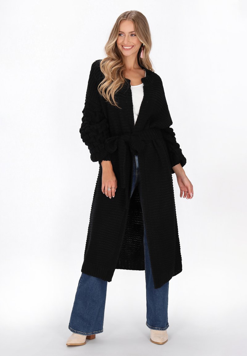 Černý pletený dlouhý cardigan s texturovanými rukávy, otevřeným předním dílem a svázaným pasem. Nesený přes bílé tričko a kombinovaný s širokými džínami.