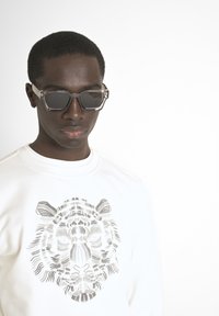Gafas de sol hexagonales grises y un sudadera blanca con un gráfico detallado de un tigre gris. El fondo es completamente blanco.