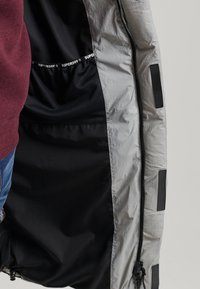 Chaqueta gris claro con forro interior negro y detalles elásticos. Cierre con cremallera en el lado derecho y paneles horizontales de tela.