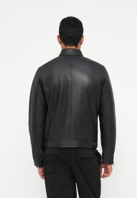 Juodas odinis švarkas su minimalistiniu dizainu, užsegamomis rankovėmis ir juostiniu apykakle. Lygus paviršius su prigludusia siluete, be matomų logotipų.