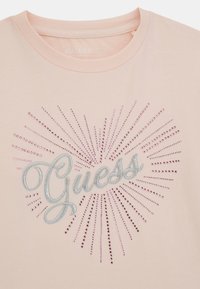 Jasnoróżowa koszulka T-shirt z organicznej bawełny z haftowanym srebrnym logo "Guess", ozdobiona różowymi kryształkami, które promieniują na zewnątrz.