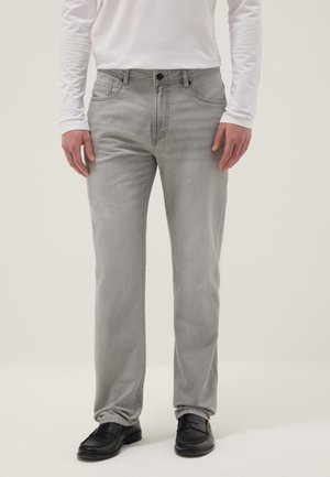 Mann trägt hellgraue Jeans mit geradem Bein, weißes Langarmshirt und schwarze Loafers, steht vor einem schlichten hellen Hintergrund.