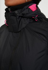 Schwarze Jacke mit hohem Kragen, ausgestattet mit einem pinkfarbenen Innenfutter und Kordelzügen. Reißverschluss und gerippte Bündchen sind sichtbar.