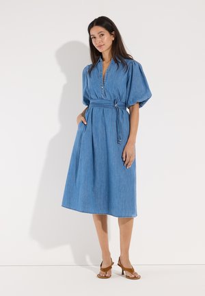 Frau trägt ein blaues Denim-Midikleid mit Puffärmeln und einem Taillengürtel, steht vor einem einfachen weißen Hintergrund mit braunen Sandaletten mit Absatz.