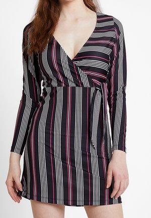 Vrouw draagt een zwarte wikkeljurk met verticale witte en roze strepen, lange mouwen en een strikkoordje in de taille.