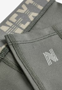 Grøn atletisk stof med en kontrastfarvet beige talje, der har et geometrisk mønster. Inkluderer syede kanter og et logo i hvidt.