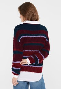 Strickpullover in Burgunder mit marineblauen, lilafarbenen und kastanienbraunen Streifen. Verfügt über eine lockere Passform, einen Rundhalsausschnitt und gerippte Bündchen. Über einem weißen Shirt getragen.