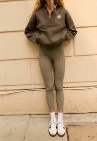 Olijfgroene zip-up hoodie met een voorzak en logo, gecombineerd met lichtgrijze leggings en witte sneakers met donkere accenten.