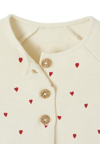 Cremfarbendes Kleidungsstück mit kleinen roten Herzmotiven und drei beigen Knöpfen am vorderen Halsausschnitt.