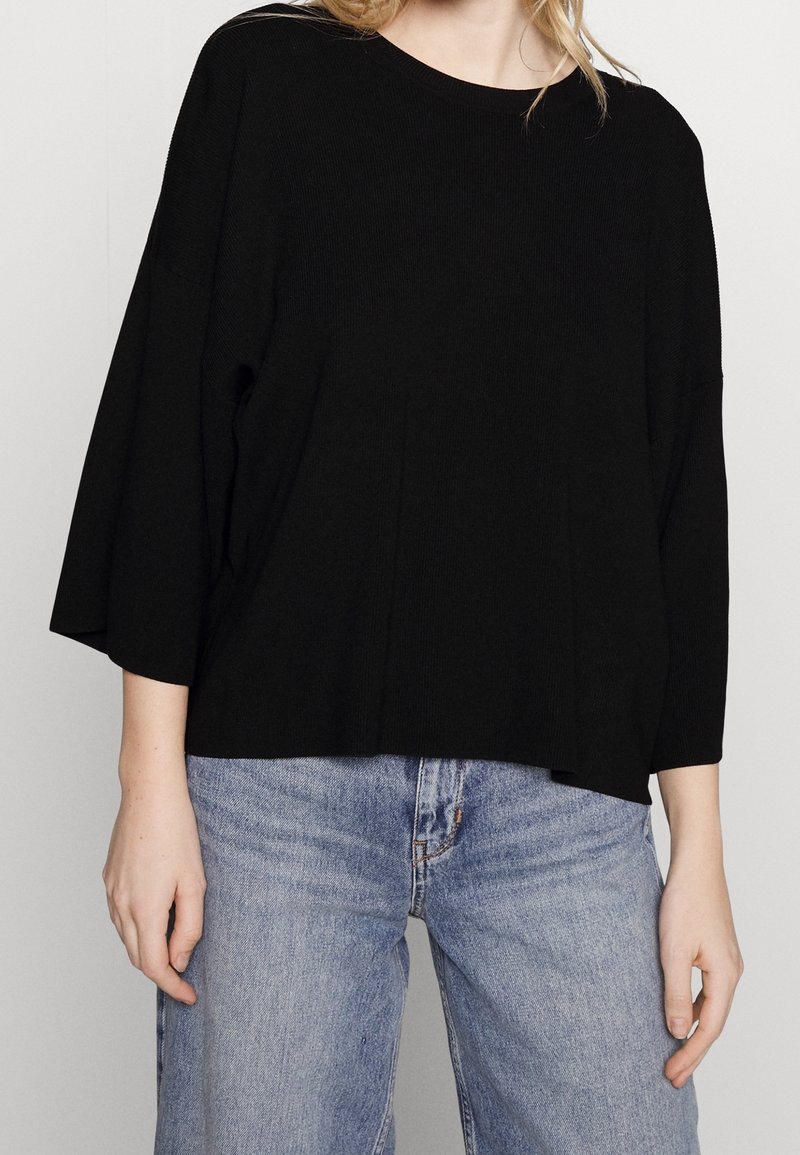 Czarna, oversize'owa bluzka z długim rękawem o prążkowanej fakturze i szerokim dekoltem, zestawiona z jasnoniebieskimi, luźno dopasowanymi jeansami.