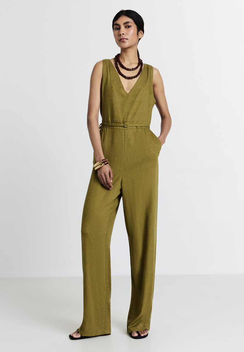 Donna che indossa un jumpsuit senza maniche verde oliva con cintura, collane in legno a strati, braccialetti e sandali neri con apertura sulla punta, in piedi con una mano in tasca.