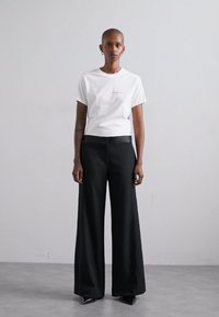 T-shirt blanc court avec un branding minimal, associé à un pantalon noir à jambes larges. Le modèle porte des talons aiguilles noirs, sur un fond neutre.