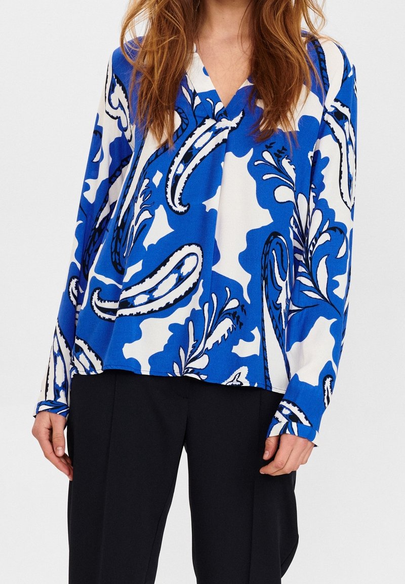 Femme portant un chemisier à manches longues bleu et blanc à motif cachemire avec un col en V, associé à un pantalon noir, debout sur un fond uni.