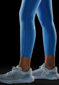 Blauwe sportlegging met een soepele textuur, voorzien van gepunte reflecterende accenten aan de zijkant. Gecombineerd met lichtgrijze, ademende sneakers.