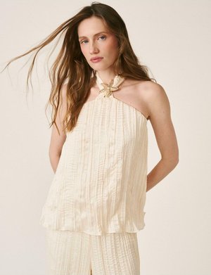 Femme aux longs cheveux portant un haut crème sans manches plissé avec une broche en forme d'étoile de mer au col, pantalon assorti plissé, posant devant un fond neutre.