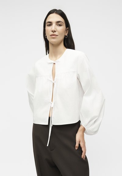 Γυναίκα με μακριά σκούρα μαλλιά φοράει λευκό μπλουζάκι με μανίκια μπαλόνι και δεσίματα, σε συνδυασμό με φαρδιά μαύρα παντελόνια σε απλό φόντο.