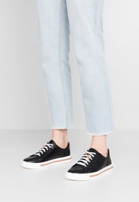 Baskets en cuir noir avec des lacets blancs, des accents contrastants en tan et une semelle texturée. Associées à un jean en denim bleu clair, cropped.