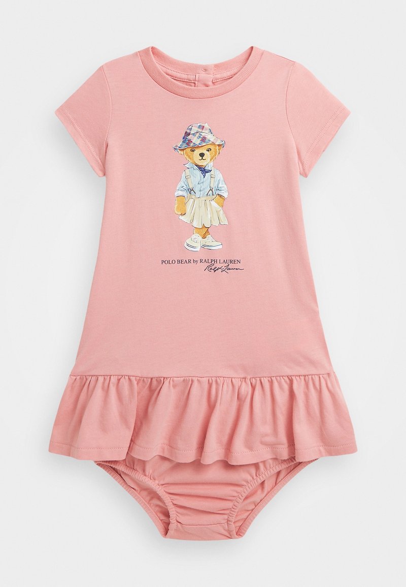 Růžové bavlněné šaty s volánkovým lemem, na nichž je obrázek medvěda v modré košili a klobouku. Krátké rukávy, měkká textura a elastický spodní lem.