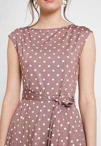 Ruskea pilkullinen mekko, jossa on hihattomat hihat ja sidottava vyötärö. Sisältää valkoisia pisteitä, sileän kankaan tekstuurin ja yksinkertaisen, tyylikkään muotoilun.