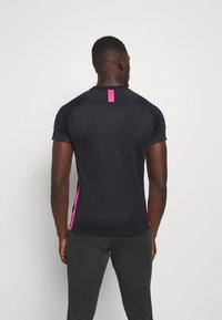 Sort atletisk polo med korte ærmer, prydet med lyserøde sideaccentuer og et vertikalt lyserødt bånd på ryggen. Fremstillet af tekstiler med struktur og åndbarhed.