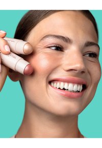 Deux bâtons de maquillage crème à double embout sont maintenus à côté d'un visage souriant. Les bâtons présentent une couleur beige et une couleur rosée, avec un design élégant et une texture légère.