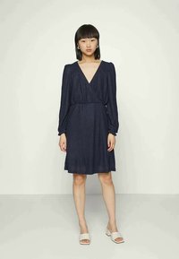 Vestido azul-marinho tipo envoltório com mangas longas e bufantes e uma cintura ajustada. Tecido texturizado, decote em v e saia rodada. Sandálias simples em bege.