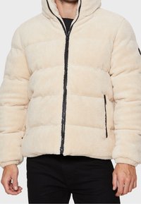 Chaqueta de abrigo de forro polar beige con cremallera frontal, capucha y detalles en negro. Presenta un acolchado horizontal y bolsillos laterales. Textura suave.