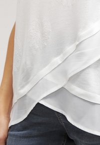 Top bianco caratterizzato da un design a strati con sovrapposizione trasparente, modelli di ricamo geometrico e una texture morbida. Abbinato a jeans scuri.