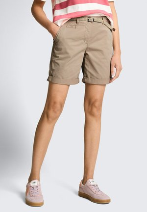 Model draagt beige opgerolde shorts met geweven riem, roze-wit gestreept overhemd en roze sneakers met rubberen zolen.