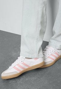Zapatillas blancas con rayas rosas, con una suela texturizada beige y una parte superior de cuero suave. Combinadas con unos pantalones de mezclilla azul claro.