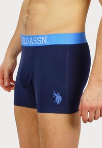 Marinefarbene Boxershorts mit einem hellblauen elastischen Bund, der mit "POLO ASSN." beschriftet ist, sowie einem passenden, auf die linke Beinseite gestickten Logo. Material aus Baumwollmischung.