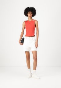 Rotes, geripptes Tanktop mit Schulterausschnitten, kombiniert mit weißen Denim-Shorts mit ausgefransten Säumen. Das Model hält eine schwarze Clutch und trägt weiße Sneakers.