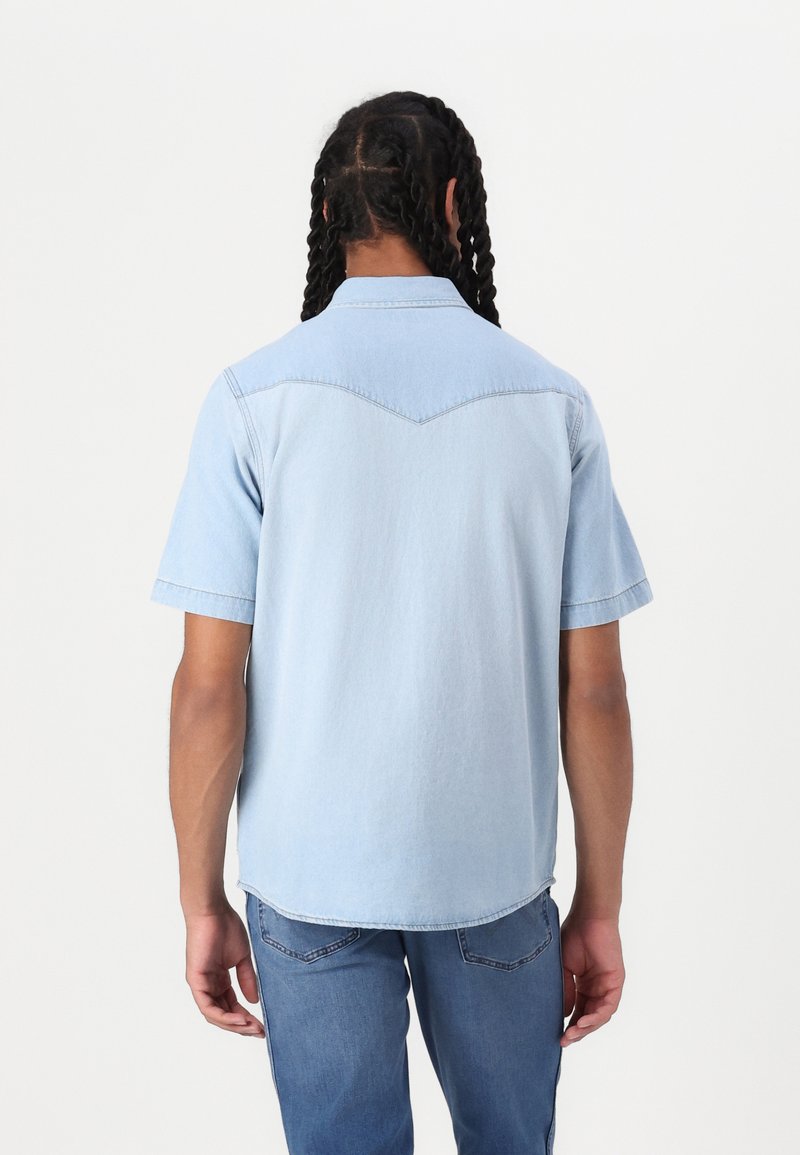 Jasnoniebieska denimowa koszula z krótkim rękawem z wyraźnym kołnierzem, widocznym szwem i zaokrąglonym dołem. Widok z tyłu pokazuje gładką teksturę i luźny krój.