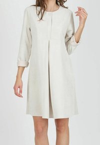 Nainen, joka on pukeutunut polvimittaisiin, pitk�ähihasiin beigekuiduista valmistettuihin mekkoihin, joissa on etujuova ja käärityt hihat, seisoo yksinkertaista taustaa vasten.