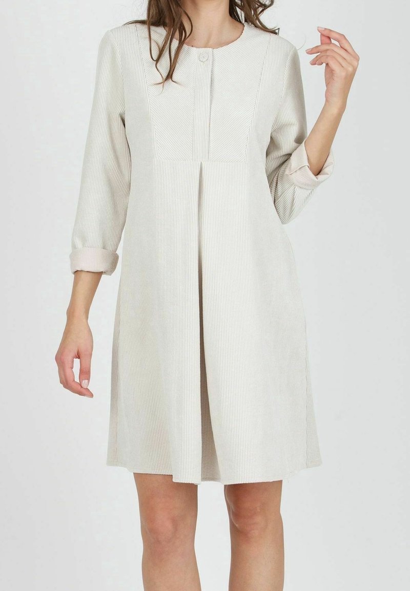 Nainen, joka on pukeutunut polvimittaisiin, pitkähihasiin beigekuiduista valmistettuihin mekkoihin, joissa on etujuova ja käärityt hihat, seisoo yksinkertaista taustaa vasten.